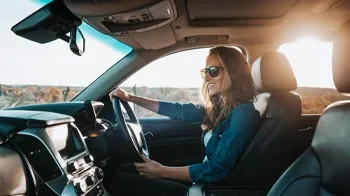 Behind the steering wheel, a smiling woman in a blue blouse keeps her eyes on the road.